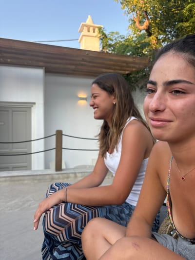 Two young adult females are seated outdoors in Platys Gialos, Greece, in what appears to be a resort or hotel common area during late afternoon. The woman in the immediate foreground, positioned to the right, is partially visible from the chest up and to the upper leg. She has dark hair, a slight smile on her visible right facial profile, and wears a multi-colored strappy top with a gold-colored chain necklace featuring a small red clover pendant. A red abrasion is present on her right knee/upper thigh. Behind her, slightly to the left, a second young adult female is seen in profile, smiling with teeth visible. She has brown hair and wears a white ribbed tank top and blue and brown geometrically patterned wide-leg pants. Her forearms rest on her knees.
The background features a white, minimalist building with a light grey door marked with the number "103" on a small plaque. A double-bar wall sconce emits warm light on the building's facade. In the mid-ground, a low barrier or railing composed of two dark vertical posts and two horizontal dark ropes separates the seating area from the building. Overhead, wooden beams form a pergola structure. Beyond the building, a light-colored, ornate tower or chimney is visible against a clear blue sky, flanked by green foliage. The ground beneath the individuals is a flat, light grey paved surface.