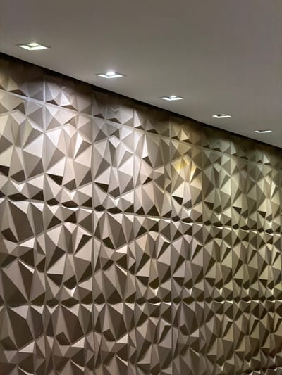 A sharp angle view focuses on two surfaces. The left is a smooth, white wall punctuated by evenly spaced, square inset lights. The right is a textured wall with a geometric pattern of triangular and polygonal shapes. The lighting from the inset fixtures casts shadows and highlights across the textured surface, emphasizing its depth and form. The color palette is predominantly neutral, with shades of white, gray, and subtle metallic tones.