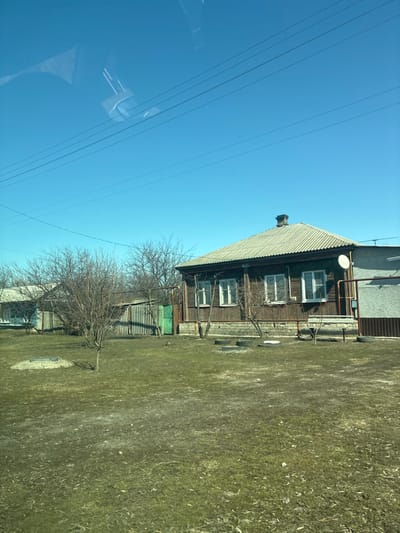 The image depicts a residential scene in Povorino, Russia, under a clear, bright blue sky with no visible clouds. The foreground consists of patchy, dried green and brown grass interspersed with bare dirt. Several dark, circular objects, resembling tires or manhole covers, are arranged on the ground in front of the main structure. A light grey, rectangular concrete slab is partially visible in the lower-left foreground.
The central subject is a single-story house with dark brown wooden siding and a light grey corrugated roof, featuring a dark brick chimney at its peak. Four white-framed windows are visible on the front facade, and a white house number sign, possibly displaying "24" or "74," is affixed above the second window from the left. The house sits on a light-colored foundation. On the right side of the house, a white circular satellite dish is mounted on a light brown metal pipe structure, adjacent to yellow gas pipes that run along the exterior and connect to a meter box. Two small, bare deciduous trees stand directly in front of the house.
To the left of the main house, a silver-grey corrugated metal fence runs horizontally, with a bright green metal gate. Behind this fence and the bare trees, a portion of another single-story house, painted light blue, is visible. Multiple parallel utility wires stretch across the upper portion of the clear blue sky. Reflections from an interior space, possibly a vehicle's dashboard or window frame, are present in the upper-left quadrant, indicating the image was captured from inside a conveyance. No individuals or active interactions are visible in the scene.