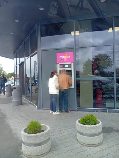 In Kuršėnai, Lithuania, two individuals are observed near a "Medus ATM" situated outside a building with a modern glass facade. A man in a brown jacket and blue jeans is actively using the ATM, his right hand near the card slot. To his left, a woman in a white puffer vest and blue jeans stands with her arms behind her back, facing the ATM. The ATM itself is mounted on a grey wall, displaying the "Medus" logo in pink and the word "ATM" above. Below the logo, "IŠDAVIMAS" is visible. Several logos of financial institutions like "Citadelė", "Swedbank", "LKU", and "Luminor" are present on the ATM interface. The ground is paved with grey rectangular tiles. In the foreground, two circular concrete planters each contain a small green shrub. Reflections in the glass show trees and possibly other buildings. In the background, two grey cylindrical trash receptacles are positioned near the entrance. Further back, several cars are parked.