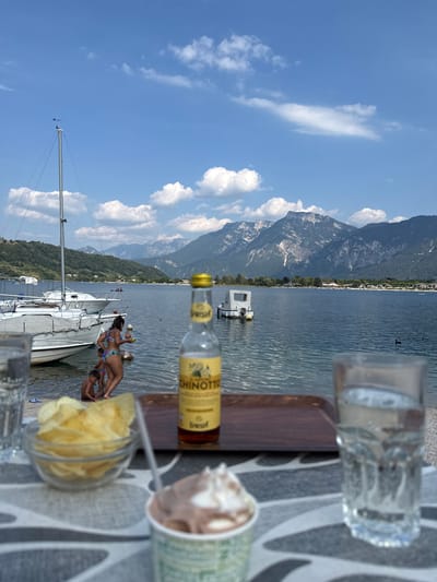 In Calceranica al Lago, Italy, a scenic view of a lake is captured on a sunny day with a beverage and snacks in the foreground. The lake is calm, reflecting the blue sky dotted with fluffy white clouds. In the distance, majestic mountains form a dramatic backdrop, their peaks partially shrouded in haze. Several sailboats are moored along the shore, their white sails contrasting with the blue water.
On the lakeside, a woman in a bikini is interacting with two children who are wading in the shallow water. The woman is holding a small object, possibly a toy, while the children are playing near the shore.
In the immediate foreground, a table is set with a glass of water, a bowl of potato chips, a bottle of Chinotto, and a cup of ice cream. The table is covered with a patterned cloth, suggesting a relaxed outdoor dining setting. The overall atmosphere is one of leisure and enjoyment, typical of a summer day by the lake.