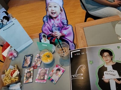A bird's-eye view captures a collection of K-pop merchandise and food items arranged on a grey surface, likely a table. In the foreground, a small cardboard container holds a portion of french fries, accompanied by a split container of ketchup and mayonnaise. To the right of the fries, a glass with yellow liquid and mint leaves sits next to a clear plastic cup with a straw, containing a beverage. Several small, rectangular items with photographs of young men adorn the table, some with colorful borders and keychains. A larger, life-sized cardboard cutout of a baby in traditional Korean attire is visible in the background, alongside another cutout of a young man. A framed poster with a picture of a man holding a "HAPPY BIRTHDAY" cake and a "REC 06:13" overlay is on the far right. The overall scene suggests a fan event or gathering related to K-pop. The city of Serang, Indonesia, is not discernible from the provided image.