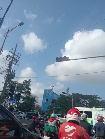 This is a vertical, eye-level shot of a busy street scene in Surabaya, East Java, on a sunny day with a partly cloudy sky. The foreground is dominated by the back of a black car, with a red motorcycle rider wearing a red and white helmet in the immediate right foreground. This rider is wearing a red jacket with a large white "ACI" logo on the back. Further back, several other motorcycle riders are visible, also wearing helmets. One rider is wearing a green helmet, another a white helmet with black and green details, and another a red helmet. A white delivery van with green and white branding that reads "PARAHITA TEMAN SEHAT" is also in the mid-ground.

The background features a blue building with multiple windows and visible telecommunication towers on top. To the left of the blue building, a green minaret of a mosque is visible above trees. Traffic lights are prominently displayed, with the red light illuminated. A street sign in Indonesian reads "Belok Kiri (Ikuti Isyarat Lampu)", meaning "Turn Left (Follow Traffic Signal)". The overall atmosphere suggests a typical urban commute with a mix of vehicles and pedestrians, likely during daylight hours due to the bright sunlight and clear sky. The presence of delivery vehicles and riders indicates ongoing commercial activity.