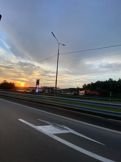 A sunset illuminates a highway in Mamony, Russia. The sky is a mix of blue and grey clouds, with the orange sun setting on the left side of the frame. A roadside billboard with colorful advertisements stands near a gas station. Cars and trucks are visible on the highway, some of which are at the gas station. A tall streetlamp stands on the right side of the road. The highway is paved with asphalt and has white lane markings. A metal guardrail separates the highway from the surrounding greenery.