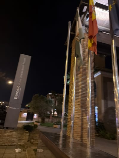 The image shows a nighttime scene in Punta Umbría, Spain. The primary subject is the "Barceló Punta Umbría Beach Resort" sign. The background features buildings, trees, and a dimly lit street. Flags are visible above, including the flag of Spain. The photograph appears to be taken from a low angle, looking upwards towards the resort signage.