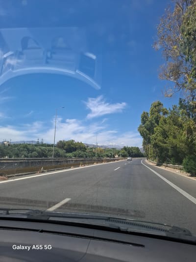 The image depicts a view from inside a moving vehicle, likely a car, traveling on a paved road in Gazi Municipal Unit, Greece. The vehicle's interior elements, such as part of the dashboard, a wiper blade, and a side mirror reflection are visible.

The road is lined with greenery, including trees and shrubs. Beyond the vegetation, the background shows the landscape with some distant features which seem to be mountains. Street lighting poles run parallel to the road.

The sky is blue with wispy clouds. The overall impression is one of a daytime shot taken while driving in a rural or suburban setting within the Gazi Municipal Unit. A phone's watermark is present at the bottom of the image which reads "Galaxy A55 5G".