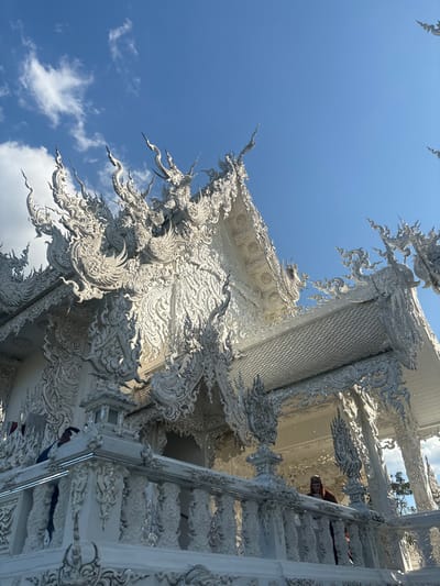 An upward-angle photograph captures a section of Wat Rong Khun, the White Temple, located in Ban Rong Khun, Thailand. The primary subject is the highly ornate, multi-tiered white temple structure, detailed with intricate sculptural elements including Naga figures, chofahs, and flame-like motifs that glisten under direct sunlight due to embedded reflective materials. The sky above is clear and blue with scattered white clouds. In the foreground, a white, elaborately carved balustrade is visible. Two individuals are present behind this balustrade: one on the left, largely obscured by a pillar, wearing a dark blue garment; and another on the right, partially visible, wearing a reddish-brown garment. Both individuals appear to be observing the temple grounds.