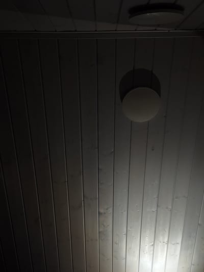 The image shows a dark wooden ceiling with two circular light fixtures. One fixture is on the upper left and is mostly in shadow, appearing as a dark circle with a faint highlight. The second fixture is on the right side of the image, also circular and illuminated, casting a bright light downwards. The ceiling planks are oriented vertically from the perspective of the image. The overall lighting is dim, with the exception of the area directly under the illuminated fixture. The location provided, Doma 1096 Km, Russia, is a postal code and likely refers to a remote or rural area, potentially a dacha or a standalone building. There are no people or visible actions depicted in the image.
