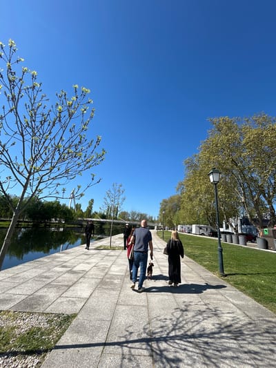 A sidewalk in Chaves, Portugal, is lined with square pavers alongside a reflective body of water. Two people are walking on the sidewalk, a man in a blue shirt and jeans walking a small brown dog, followed by a woman with long blonde hair wearing a dark coat. Tree shadows stretch across the sidewalk. In the background, a row of RVs is parked along the grass. A blue sky with a few scattered trees is overhead.