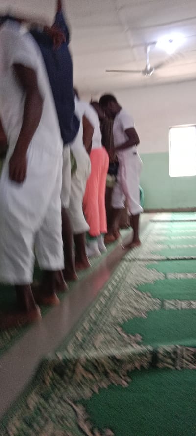 The scene depicts an indoor gathering of at least six individuals in the city of Iyara, Nigeria. The room features light-colored walls, with the lower section painted a light green and the upper section off-white. The floor is covered with green patterned carpets or prayer rugs. A bright rectangular window is visible on the right wall, providing natural light. A silver-colored ceiling fan and a luminous light fixture are present on the off-white ceiling.
The individuals are positioned in a linear formation, primarily facing away from the camera, with their feet aligned along a subtle raised line or demarcation on the carpet. Most individuals are wearing loose-fitting white clothing. One person is dressed in dark blue attire, and another is wearing bright pink/coral trousers. Individuals appear to be barefoot or wearing very light footwear. No specific actions or interactions between the individuals are clearly discernible beyond their static alignment. The overall lighting is bright, suggesting adequate illumination from both natural and artificial sources.
