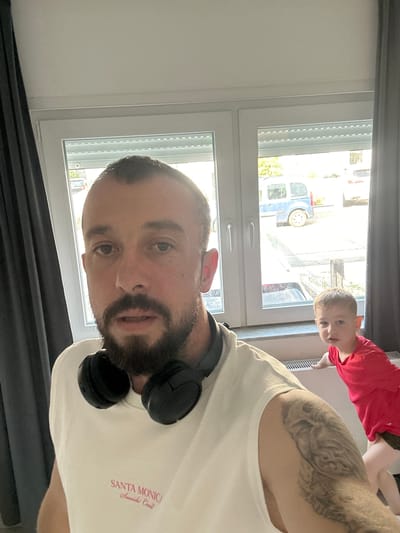 A man with a beard and headphones around his neck is taking a selfie in front of a window. He is wearing a white t-shirt with "SANTA MONICA Seaside Coast" written on it. Behind him, a young child in a red t-shirt is partially visible near a radiator. The window looks out onto a street with a blue van and other vehicles, suggesting an urban or suburban setting, potentially in Schwetzingen, Germany. The lighting indicates it is daytime.