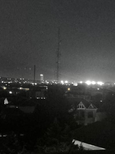 This is a black and white, slightly grainy photograph taken at night, likely from an elevated position, overlooking a city. The foreground is dominated by dark silhouettes of trees and foliage, obscuring much of what lies beneath. In the midground, there is a distinct, two-story house with a prominent gable roof and visible windows. Beyond this house, the city stretches out, characterized by a multitude of lights.
The city lights are varied; some are small and scattered, suggesting residential areas or streetlights, while others are larger and brighter, indicative of commercial buildings or major thoroughfares. A tall, slender radio tower or transmission mast is visible piercing the skyline, a prominent vertical element against the horizontal spread of the city. In the far distance, a tall, illuminated building stands out, its lit windows forming a bright rectangle against the dark sky, a possible landmark or skyscraper.
The overall scene suggests an urban environment at night. The sky is dark, with no visible stars or moon, and the city lights are the primary source of illumination. There are no visible people. The presence of trees in the foreground and the scattered nature of the city lights might imply a location on the outskirts of the city, looking inwards. The time of day is clearly night, and the weather appears clear, with no indications of rain or fog. No text is visible in the image. The mood is somewhat subdued due to the low light conditions and the black and white processing, giving it a slightly stark or moody aesthetic. The location is identified as Bina, Azerbaijan.