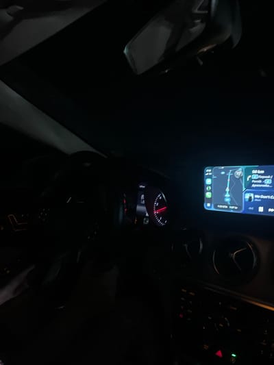 The image appears to be taken from inside a car at night. The dashboard is visible, with a phone displaying a navigation app mounted on it. The app shows a route indicating 56km to a location named "Перник" via routes numbered 116 and 117. The ETA on the navigation screen is 1:23 for an arrival time of 1:07. The speedometer needle is barely visible, but it shows a low speed. To the right of the phone are air vents and buttons of the car's infotainment system. The environment outside is dark, and the car is on a road or street. The location is in relation to the city of Pernik and a destination of “Anaaunolo”, both likely near Mursalevo, Bulgaria, as that is the implied current location. There are no visible people or interactions taking place.