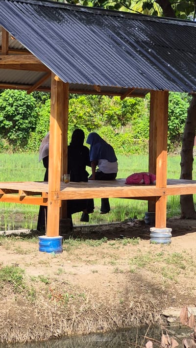 The image depicts a scene under a corrugated metal roof structure in Raya Sanggeu, Indonesia. Inside the structure, three individuals are present. One person is kneeling, wearing a blue shirt with a white undershirt visible. Another figure, entirely in black, stands behind the kneeling person. The third individual, partially obscured, is wearing a white head covering. On a wooden beam above, a pink stuffed animal is visible. The structure is supported by wooden posts secured with blue and grey plastic fittings at the base. The surrounding environment consists of lush green vegetation and a patch of dry grass and soil.