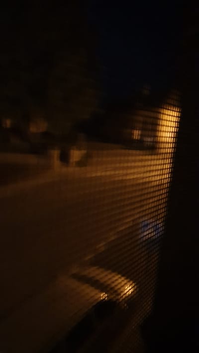 The image shows a blurred view of a street at night, seen through a screen or mesh. The street is illuminated by streetlights or lights from buildings, casting a warm, orange glow. There are faint shapes of buildings or houses visible in the background, but they are indistinct due to the low light and blurriness. The foreground is dominated by the pattern of the screen, which creates a grid-like effect over the scene. The overall impression is one of looking out a window at a quiet, dimly lit street. The location is identified as Vrchotovy Janovice, Czechia.