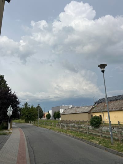 The image shows a street scene in Hulín, Czechia, captured on a cloudy day. The perspective is from a low angle, looking upwards towards a large, overcast sky filled with dramatic, textured clouds. A streetlamp with a distinctive, round fixture stands prominently in the foreground, casting a stark silhouette against the sky.
To the right of the streetlamp, a building with a red-tiled roof and yellow siding is visible, suggesting a residential or commercial area. A paved road curves into the distance, bordered by a sidewalk constructed with light-colored paving stones and a decorative red brick inlay. Grassy areas with sparse vegetation are situated between the sidewalk and the building. Further down the street, more trees and buildings can be seen, with some featuring gray, modern facades. A directional sign with two white arrows pointing left is positioned near a cluster of dark foliage. The overall impression is that of a quiet, perhaps residential, street under a sky that suggests impending rain or a change in weather. The time of day appears to be daytime, likely late afternoon given the light. No people are visible in the frame.
