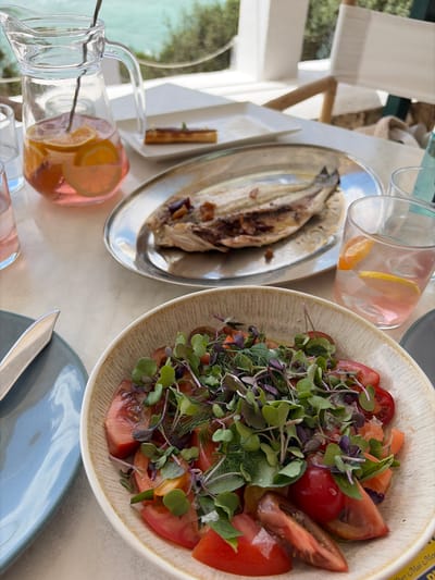 This image captures a close-up, overhead view of a dining table set for a meal, likely outdoors given the bright lighting and hints of greenery in the background. The focal point is a meal consisting of a whole roasted fish served on a silver platter and a vibrant salad in a textured, off-white bowl. The salad is a colorful mix of chopped tomatoes, microgreens, and dill. Adjacent to the food are two glass carafes filled with a pinkish liquid and fruit slices, possibly infused water or a light beverage.
The scene suggests a leisurely meal, perhaps lunch or an early dinner, on a clear day. The bright lighting and the casual presentation imply a relaxed atmosphere. The presence of the fish and salad, along with the refreshing drinks, points towards a meal that is both fresh and flavorful. The table appears to be made of a light-colored material, possibly marble or a similar stone, and is placed in an area with some dappled sunlight filtering through.
Notable details include the glistening texture of the roasted fish, the fresh appearance of the salad ingredients, and the condensation on the glass carafes, indicating they are chilled. There are no people visible in the frame, focusing the attention entirely on the food and dining setup. The overall impression is one of a well-prepared and inviting meal in a pleasant setting.