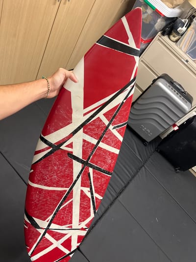 A human hand and forearm, adorned with a green and dark-colored braided bracelet, holds a red, white, and black striped surfboard vertically. The surfboard exhibits extensive wax residue and surface wear. The scene is an indoor environment located in Rancho Palos Verdes, United States. In the background to the left are light-colored wooden cabinet panels. To the right, a storage area is visible, featuring cluttered open shelves at the top, a silver-grey hardshell rolling suitcase with a wavy texture in the mid-ground, and a partially obscured black hardshell rolling suitcase beneath it. The floor is covered with dark grey padded mats showing distinct seams. No other individuals or interactions are present. The lighting is uniform, consistent with an indoor setting.
