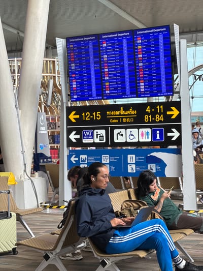 The image depicts an interior view of an international departure terminal at Phuket International Airport in ตำบล ไม้ขาว, Thailand. In the foreground, two individuals are seated on light brown, wicker-style airport benches with white metal frames. On the left, a male with light-brown skin, dark hair tied back, and glasses, wearing a dark blue long-sleeved jacket, bright blue Adidas track pants with white stripes, white "VANS" socks, and black sneakers, is operating a silver laptop on his lap. He has white wireless earbuds in his ears. To his right, a female with dark hair, wearing a dark green t-shirt and brown patterned pants, is holding and looking at a bright yellow smartphone. Her left forearm displays tattoos. A light green wheeled suitcase is partially visible on the far left next to the seating.
In the midground, two large, blue digital "International Departure" screens display flight information, including destinations such as Mumbai, Kolkata, Bengaluru, Perth, Singapore, and Suvarnabhumi, with the date "06 Jun 2026" visible. Below these screens, a prominent yellow and black overhead sign directs passengers, indicating "Gates | 登机口" (Gates) with arrows pointing left for gates "12-15" and right for gates "4-11" and "81-84". Below this, a white and black sign features arrows pointing left for "VAT REFUND FOR TOURISTS" and "DUTY FREE", and right for symbols indicating accessible restrooms, male/female restrooms, and an information desk. A lower blue sign provides icons for "Currency Exchange," "Food & Beverage," "Observation Deck," and "CIP Lounge," with text in Thai, English, and Chinese/Russian.
The background reveals typical airport architecture including towering white cylindrical support columns, metal structural elements, and a "FREE CHARGER" station with the "AOT" (Airports of Thailand) logo. Shelves stocked with duty-free items are visible behind the charging station. Other passengers are scattered throughout the concourse, some seated and some standing, in an uncrowded but active environment. The floor is covered with patterned tiles in shades of brown, grey, and white, and the terminal is well-lit.