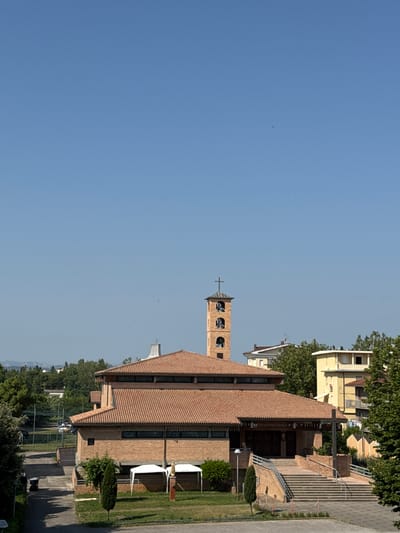 The image shows a brick church with a tiled roof and a bell tower. The church is located in Rimini, Italy, and is situated in a suburban area with trees and other buildings in the background. There are no people visible in the image. The scene is a daytime view under a clear blue sky.