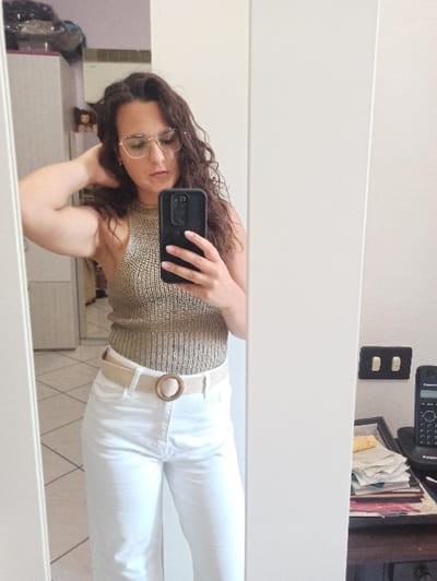 An indoor scene, presumably located in San Marzano sul Sarno, Italy, is captured via a full-length mirror selfie.
A female individual, appearing to be in her twenties or thirties, stands centered in the reflection, facing the mirror. She has long, dark, wavy hair and wears gold-rimmed eyeglasses. Her attire consists of a sleeveless, ribbed, light brown or olive-green knit top and high-waisted white wide-leg trousers, cinched with a beige woven belt featuring a round wooden buckle. Her left hand is raised, with fingers resting on the back of her head/hair, while her right hand holds a black smartphone, positioned to take the mirror selfie. The subject's gaze is directed downwards.
The environment reflected in the mirror includes light-colored, possibly cream or beige, square floor tiles arranged diagonally. To the subject's left, a white modular cabinet or wardrobe unit is visible, with various items, including what appears to be clothing or bags, stacked on its top surface. Outside the right edge of the mirror frame, a dark wooden surface (likely a desk or table) holds a stack of papers or magazines, a black Panasonic landline telephone, and a black electrical outlet mounted on a light-colored wall above the surface.