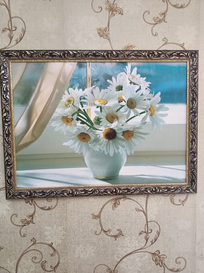 A framed still life painting of daisies in a white vase is displayed on a wall covered with light beige wallpaper featuring a repeating brown floral pattern. The painting depicts a bouquet of white daisies with yellow centers, arranged in a white vase. The vase sits on a white surface, likely a windowsill, which is illuminated by sunlight casting distinct shadows. Behind the vase, a window is visible, partially obscured by a sheer, light-colored curtain. The background outside the window appears to be a bright blue sky or water. The painting is framed by an ornate, dark brown and gold border. The overall scene is a still, domestic interior. The specific location within the city of Kürşım, Kazakhstan, is not discernible from the image.