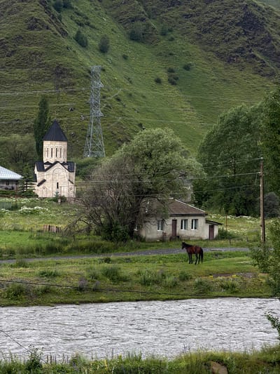 The image depicts a rural landscape with a church, a house, a horse, a river, and a power line tower. The scene is set against a backdrop of lush green, terraced hills. In the foreground, a wide, turbulent river flows, its surface rippled with movement. The riverbank is lined with vegetation, including tall grasses and wildflowers.
To the left, a stone church with a dark, conical roof stands prominently. Its architecture features arched windows and decorative stonework. Adjacent to the church, a simpler, white-roofed building can be seen.
In the middle ground, a small, weathered house with a grey roof and white-trimmed windows is situated. In front of this house, a dark brown horse is grazing on the grassy ground, wearing a reddish-brown blanket or covering.
A large, leafy tree stands between the church and the house, its branches partially obscuring the view. A tall, lattice-like power line tower is visible on the hillside behind the church, with power lines stretching across the frame. The hills themselves are covered in thick, green grass and dotted with small trees and shrubs. The sky appears overcast, contributing to the subdued lighting of the scene. The image captures a serene, yet somewhat austere, view of rural life in the depicted location.