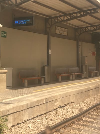 The image depicts a train station platform in Montcada i Reixac, Spain. A digital sign displays "R4 TERRASSA EST DEL NORD". To the left, a green sign with an arrow indicates direction. The platform features several metal benches with orange legs, some of which are empty, while others are occupied by people out of frame. Two cylindrical trash receptacles are visible. In the background, a wall is partially obscured by the train station's canopy, supported by metal beams. The platform itself is made of concrete with yellow safety lines and is bordered by gravel. Further down the platform, more benches and a vending machine are present. The overall environment suggests a functional, public transportation hub.