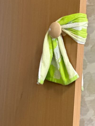 A close-up shot shows a light brown wooden door with a towel hanging on a wooden hook. The towel is patterned with green and white stripes and has a fuzzy texture. The hook is round and light brown, and it is attached to the edge of the door. The background is a wall with a textured pattern. The image is well-lit and the focus is on the towel and hook. The scene is simple and domestic.