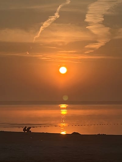 The image depicts a sunset over a body of water, possibly a sea or a large lake, near the city of Surakovo, Russia. The sun, a bright orange orb, is positioned slightly above the horizon, casting a warm glow across the sky. The sky is filled with a mix of orange and yellow hues, with white contrails from an aircraft creating abstract patterns. In the foreground, a dark silhouette of a sandy shore is visible. Two figures, also in silhouette, are crouched on the shore, facing the water. The water reflects the vibrant colors of the sunset, creating a shimmering effect. A dotted line, possibly a marker or a boundary, is faintly visible across the water. The overall scene conveys a sense of calm and serenity.