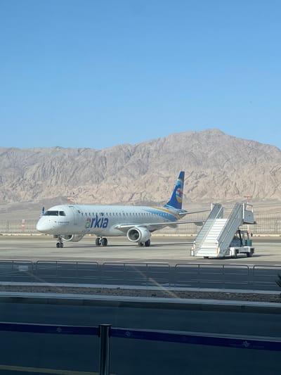 A white and blue Arkia Airlines plane is parked on the tarmac at the Ramon Airport in Be'er Ora, Israel (latitude: 29.725, longitude: 35.007). The plane is facing the camera and is connected to a mobile airstair. The plane has a blue tail with a white logo, and a white body with a blue stripe running down the side. The plane's registration number is 4X-ACB. The plane is parked in front of a set of mountains. The sky is blue and clear. There is a black fence in the foreground, with a blue and white striped ribbon attached to it. There is a line of dark bushes on the right of the plane, and a white building in the background.