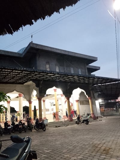 A mosque in Geulanggang Teungoh, Indonesia, is captured from a low angle in the twilight. The mosque features a dark-colored roof with a textured, thatched overhang in the foreground. The building itself is multi-leveled with a prominent dome and several arched doorways supported by large pillars. The pillars are a mix of white and yellow with reddish-brown accents at their bases and tops. A dark metal trellis-like structure extends from the mosque's upper level, providing shade. Several motorcycles are parked on the paved area in front of the mosque. Some individuals are visible near the entrance and on the steps. A street light is illuminated on the right side of the frame. The sky is a pale blue, indicating the transition from day to night.