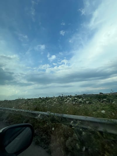 The image is a low-angle shot taken from inside a vehicle, likely while in motion, looking out towards a rural landscape. The sky occupies the top two-thirds of the frame, filled with a mix of blue sky and scattered white and grey clouds. In the foreground, a portion of the vehicle's interior is visible on the left, including a dark-colored side-view mirror. A metal guardrail runs horizontally across the middle of the image, separating the road from a grassy embankment dotted with small white flowers and green vegetation. In the background, a cluster of buildings and trees is visible, suggesting a semi-urban or suburban area beyond the immediate roadside. The overall impression is one of a journey through a moderately cloudy, daytime environment in a rural or exurban setting, possibly on the outskirts of Barletta, Italy.