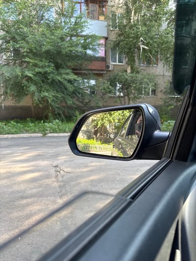An exterior shot from inside a vehicle shows a side-view mirror reflecting trees and a building. The mirror is black with a silver trim. The building is light gray with multiple windows and balconies. Lush green trees are in front of the building, with sunlight filtering through the leaves. The road in the foreground is asphalt with some cracks. The image is taken from the perspective of a person sitting in the driver's or passenger's seat of a car. The car's interior, including a black door panel and a portion of the window frame, is visible in the foreground. The scene appears to be in a residential area of Novotroitsk, Russia, likely during daylight hours given the bright sunlight and shadows.