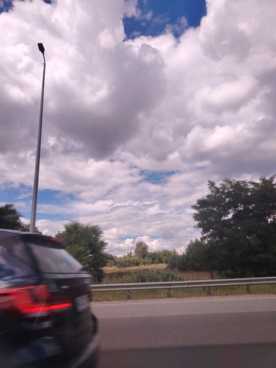 The image shows a view from a vehicle traveling on a paved road in Peremohy Street, Ukraine. A blurred dark-colored SUV is visible in the foreground, moving from left to right. The road is lined with a metal guardrail and then a grassy embankment with vegetation and trees. In the background, there is a wide expanse of sky filled with fluffy white and gray clouds against a backdrop of blue sky. A tall lamppost stands on the left side of the road, extending upwards into the sky. The overall impression is of a typical roadside scene on a partly cloudy day.