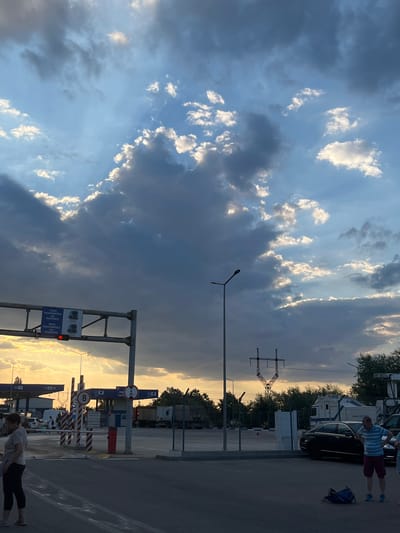 The image captures the Palanca border crossing in Moldova during a dramatic sunset. The sky is filled with a mix of dark, brooding storm clouds and bright, illuminated patches where the sun is breaking through, creating a dynamic contrast. In the foreground, the asphalt road shows lane markings.

The middle ground features the border control infrastructure, including a white overhead gantry with signs indicating "TOATE PASAPOARTELE" (All Passports) and a red traffic light. Several vehicles, including cars and trucks, are visible within the border control area, some appearing to be in queues. There are also barriers and booths typical of a border crossing. A lamppost stands prominently to the right of the gantry.

On the left side of the image, two individuals are partially visible, one a woman standing on the road, the other a person further back in the border area. On the right, two men are standing near parked cars, one in a striped shirt and shorts, looking down, and another partially visible to his right. The background is populated by trees and more border crossing structures, including power lines and a prominent lattice tower. The overall scene conveys a sense of transition and the end of a day at an international border.