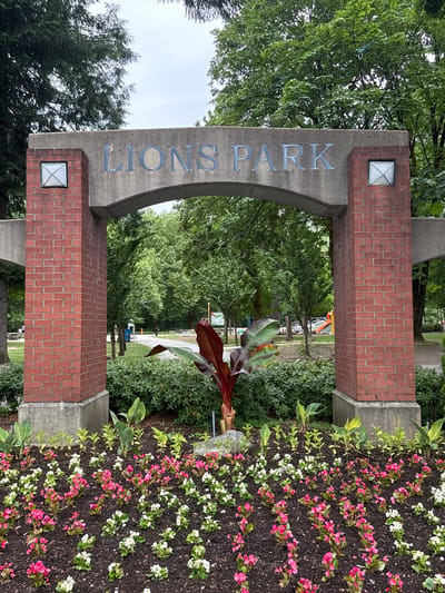 The image shows the entrance to Lions Park in Port Coquitlam, Canada. The entrance features a concrete archway supported by red brick pillars. The words "LIONS PARK" are inscribed in metal letters on the concrete arch. Two square light fixtures are mounted on the upper corners of the brick pillars. In front of the archway is a garden bed filled with blooming pink and white flowers. Behind the garden bed and archway, the park is visible, with trees, a path, picnic tables, and a playground with a slide. The sky is overcast.
