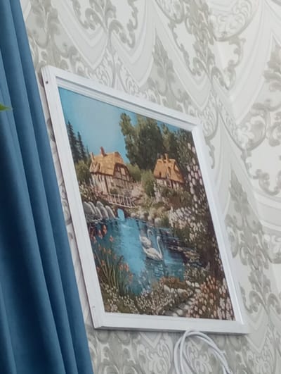 The scene, stated to be from Yaromaska, Russia, depicts an interior wall featuring a framed picture and elements of room decor. The image itself provides no visual information to corroborate its stated geographical location.

The primary subject is a rectangular picture in a white frame, mounted on a wall covered with light-toned wallpaper featuring a repeating grey ornamental or scroll pattern. The picture, which appears to be a painting or print, depicts a pastoral landscape scene. Within the artwork, two thatched-roof cottages are visible, one situated on the left and another on the right, set amongst numerous green trees. A small wooden bridge spans a calm body of blue water, where two white swans are seen swimming in the lower-middle section. The banks of the water and the foreground are densely populated with various flowering plants and rocks.

To the left of the framed picture, a segment of a vertical, pleated blue curtain is visible. Below the lower right corner of the picture frame, a coiled white electrical cable is present against the wallpaper. The photograph is captured from an oblique angle, looking slightly upwards and to the right, causing the framed picture to appear slightly skewed from a frontal perspective.
