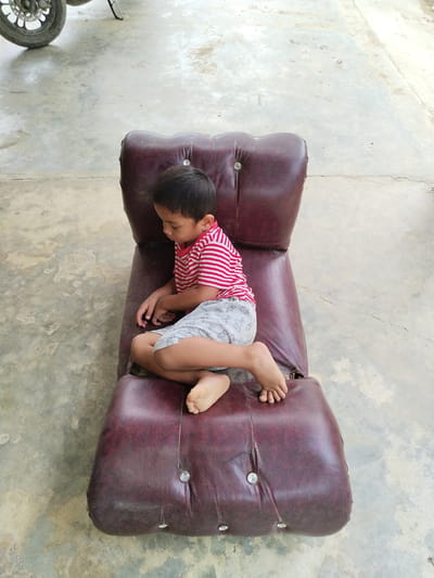 A high-angle outdoor shot in Teupinbayu, Indonesia, depicts a young boy seated on a worn, dark red tufted lounge chair positioned on a light grey concrete surface. The boy, of indeterminate age but appearing to be a toddler or young child, wears a red and white horizontally striped short-sleeve shirt and light grey shorts adorned with repeating rabbit or bunny patterns. He is barefoot, with both feet visible. His head is turned downwards, and his hands are resting in his lap, suggesting he is looking at something below him or lost in thought.

The lounge chair, made of a synthetic leather-like material, shows significant signs of wear, including cracks, discoloration, and peeling. It features circular metallic buttons, some of which are loose, missing, or attached to frayed threads, creating a tufted effect. The concrete ground exhibits variations in tone, with darker patches indicating moisture or staining. In the upper left quadrant of the image, the front wheel of a vehicle, likely a motorcycle, with a visible disc brake and part of a kickstand or frame, is partially visible. The scene is illuminated by bright, diffuse overhead light.