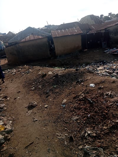 The image depicts a scene in a part of Gwagwa, Nigeria characterized by dilapidated structures and extensive refuse. Several buildings with corrugated metal roofs and earth-toned walls are visible, some appearing to be in a state of disrepair. The ground is heavily strewn with a considerable amount of garbage, including plastic waste, debris, and organic matter. A person, partially visible on the left side of the frame, is dressed in dark clothing. The overall environment suggests an area with poor waste management and substandard living conditions.