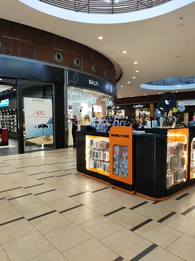 The image shows the interior of a shopping mall. In the foreground, there is a "PHONE-STORE" kiosk with a display of phone cases. To the left of the kiosk, there is a shop with a sign that reads "FATALES Summer Colors" and a discount offer of "-50% sur le 2ème produit make-up acheté". Behind this shop, there are other stores with signs like "io", "celio*", and "BALTI". A woman with blonde hair, wearing a black t-shirt and pants, is standing near the "FATALES" shop, leaning against the glass. The floor is tiled with a light-colored pattern. The ceiling has a modern design with wooden slats and lighting. The overall environment suggests a commercial retail space. Based on the identified store names and the general appearance of a modern mall, the location could be interpreted as a commercial center in Tunisia.