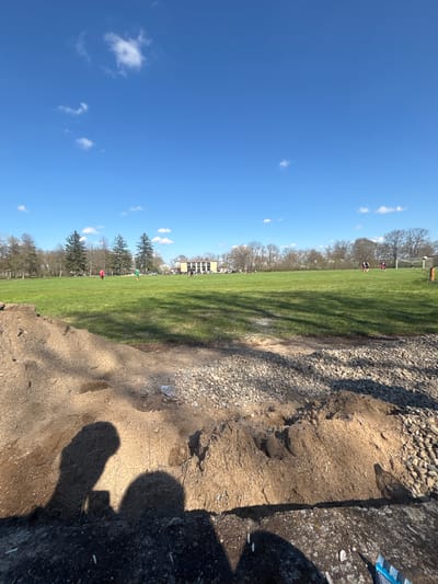 The image shows a park scene on a sunny day. A large grassy field with scattered trees and buildings in the background occupies the majority of the view. Several people, likely engaged in recreational activities, are visible on the field. The foreground features a rough terrain of dirt and rocks. The bottom right corner of the image contains a dark shadow, potentially of a person taking the photograph. There is no indication of this scene being located in Gromshin, Bulgaria.