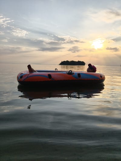 A person is in an inflatable tube on the water at sunset. The person is wearing a red cap and sunglasses, and their legs are visible sticking out of the tube. The sun is setting behind a small island in the background, casting a warm glow on the scene. The water is calm and reflects the sky and the inflatable tube. The location is the Vättern lake in Motala, Sweden.