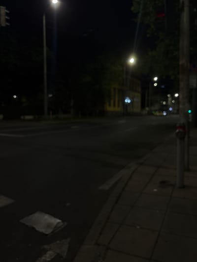 A blurry, nighttime street scene in Burgas, Bulgaria, shows a sidewalk on the right with tiled paving and a vertical pole with a red fire hydrant on it. Across the street, a yellow building is visible with illuminated windows. Streetlights are present, casting a dim glow. The road surface is dark, with faint white markings, suggesting it is a pedestrian crossing. A silver object, possibly a trash receptacle, is near the curb. Trees with green foliage are scattered throughout the background. The overall impression is a quiet, urban night.