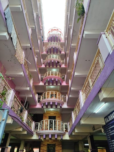 The image displays an upward-angled view within a multi-story residential building in Anantapur, India, observed from a central courtyard or atrium. The structure features at least six visible floors with a prominent central stack of successively smaller, rounded balconies. These central balconies are adorned with ornate cream-colored metal railings, pink arch-like structures, and purple horizontal bands, culminating in a dome-like element at the top, visible against an overcast sky. Flat balconies with cream-colored grid-patterned metal railings line the left and right walls.

The building's predominant colors are pink for walls and balcony undersides, a darker purple for horizontal structural bands, and cream/yellow for railings. Electrical wiring, characterized by black cables, crisscrosses the open space between floors. Various fluorescent tube lights and smaller decorative bulbs are affixed to the undersides of balconies.

Evidence of residential activity includes a grey patterned blanket and a multicolored patterned fabric draped over railings on the upper left balconies, indicating laundry. Potted plants are visible on several balconies, particularly on the lower central and right-side balconies. An external air conditioning unit is mounted on a lower-left balcony. A partial dark blue sign with white text, positioned vertically on the lower right, reads "RAJAHAM" and lists numbered entries, such as "1. 001 P. MAGABHUSHANAM" and "2. 002 S.R.NI," suggesting tenant information or unit designations. No individuals are present within the frame. The building exhibits standard concrete construction with painted finishes, showing some minor surface wear.