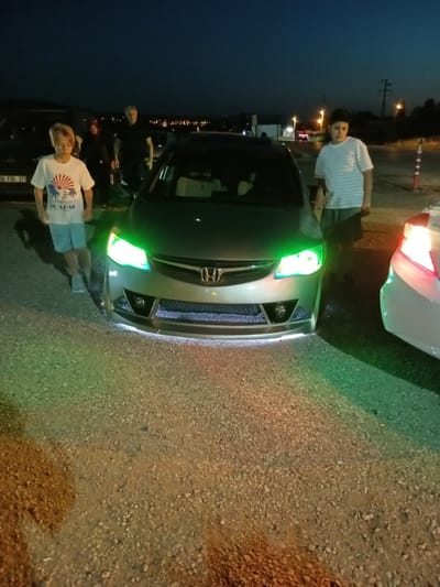 In Kırıkkale, Turkey, at night, two young boys are standing next to a silver Honda Civic that has been modified with green underglow lighting and green headlights. The car is parked on a gravel surface. The boy on the left is wearing a white t-shirt with a graphic design and light blue shorts. The boy on the right is wearing a striped t-shirt and dark shorts. In the background, other vehicles and lights from a city can be seen. The overall scene appears to be a casual gathering or car meet.