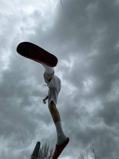 The image is a low-angle shot of a person in mid-air. The person is wearing white shorts, white socks, and red shoes. The sky is overcast and cloudy. Bare tree branches can be seen on the right side of the image. A dark object, potentially a lamppost or pole, is also visible on the right. There is no indication of this image being located in Olingen, Luxembourg.