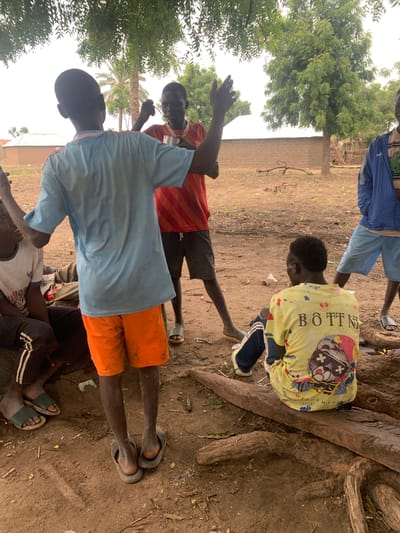 The image depicts a group of young people gathered outdoors in what appears to be a rural setting in Dull A, Nigeria. The ground is dusty earth, with some dry vegetation and tree roots visible. In the background, there is a brick wall and trees with lush green foliage, suggesting it might be a warm day with some shade from the trees.

Several individuals are visible, mostly wearing casual clothing. One person in the foreground is wearing a light blue t-shirt and bright orange shorts, with dark sandals. To their left, another person is wearing a red and white striped t-shirt and dark shorts. Further back, a person in a yellow t-shirt with a graphic print is kneeling near a large tree trunk. This yellow t-shirt has visible text that reads "BÖTTN" and some other words that are less clear. Another individual in the background is wearing blue shorts.

The people appear to be interacting, though the specific activity is not clear. Some are looking towards each other, while others are looking downwards. The overall mood seems relaxed and informal. The lighting suggests it is daytime, possibly mid-morning or afternoon, with diffused light filtering through the trees. The scene conveys a sense of community and everyday life in a Nigerian village.