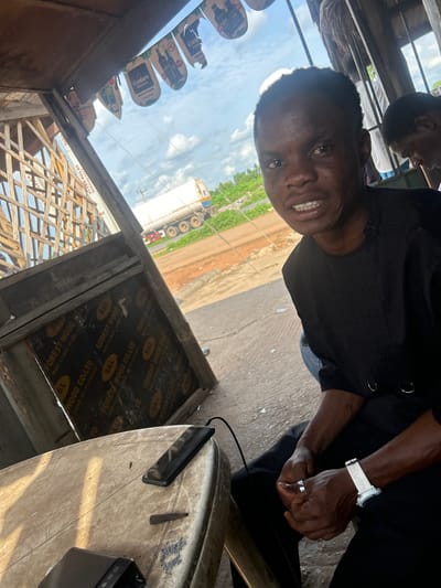 In Abobo, Nigeria, a young man with dark skin and short hair is seated at a table in an outdoor setting, possibly a roadside stall. He is wearing a black, traditional Nigerian outfit with a V-neck and decorative metal rings on the chest. His facial expression is animated, with his mouth open and teeth visible, suggesting he is speaking or laughing.
On the table in front of him are two mobile phones, one lying flat and the other partially visible at the bottom left, and a power bank. A white wristwatch with a digital display is worn on his left wrist. His hands are clasped together, and he is wearing a ring on his left ring finger.
Behind him, another person with dreadlocks is visible, and further in the background, a white tanker truck is driving on a road lined with greenery. The scene is framed by a rustic wooden structure, with what appear to be advertising banners hanging overhead, depicting bottles and text, including the brand "Cor Wam Bitters." The ground is paved with concrete. The overall atmosphere suggests a casual, everyday scene in a bustling urban environment.