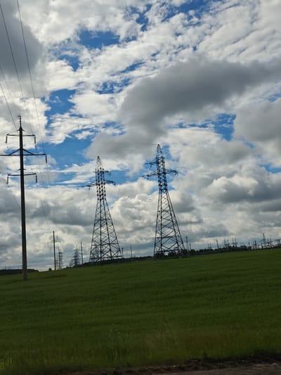 The image shows a rural landscape in the Salonski Sel'ski Sovet, Belarus, dominated by a green field and a dramatic cloudy sky. In the foreground, a lush green field stretches across the frame, with a dirt path visible at the very bottom edge.
Several power transmission towers are scattered throughout the landscape. On the left side, a single, tall, dark-colored power pole stands prominently. To its right, two larger, lattice-style power towers rise from the field, their dark metal structures contrasting with the bright sky. Further in the distance, a series of smaller power transmission towers and associated infrastructure are visible, suggesting a significant electrical substation or power line route.
The sky is filled with a mixture of bright blue sky and large, voluminous white and grey clouds, creating a dynamic and somewhat dramatic atmosphere. The lighting suggests it might be a partly cloudy day. There are no people or animals visible in the image. The scene conveys a sense of industrial infrastructure integrated into a natural, agricultural setting.